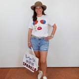 Woman wearing a white t-shirt with colorful design, denim shorts, and a hat, holding a tote bag.