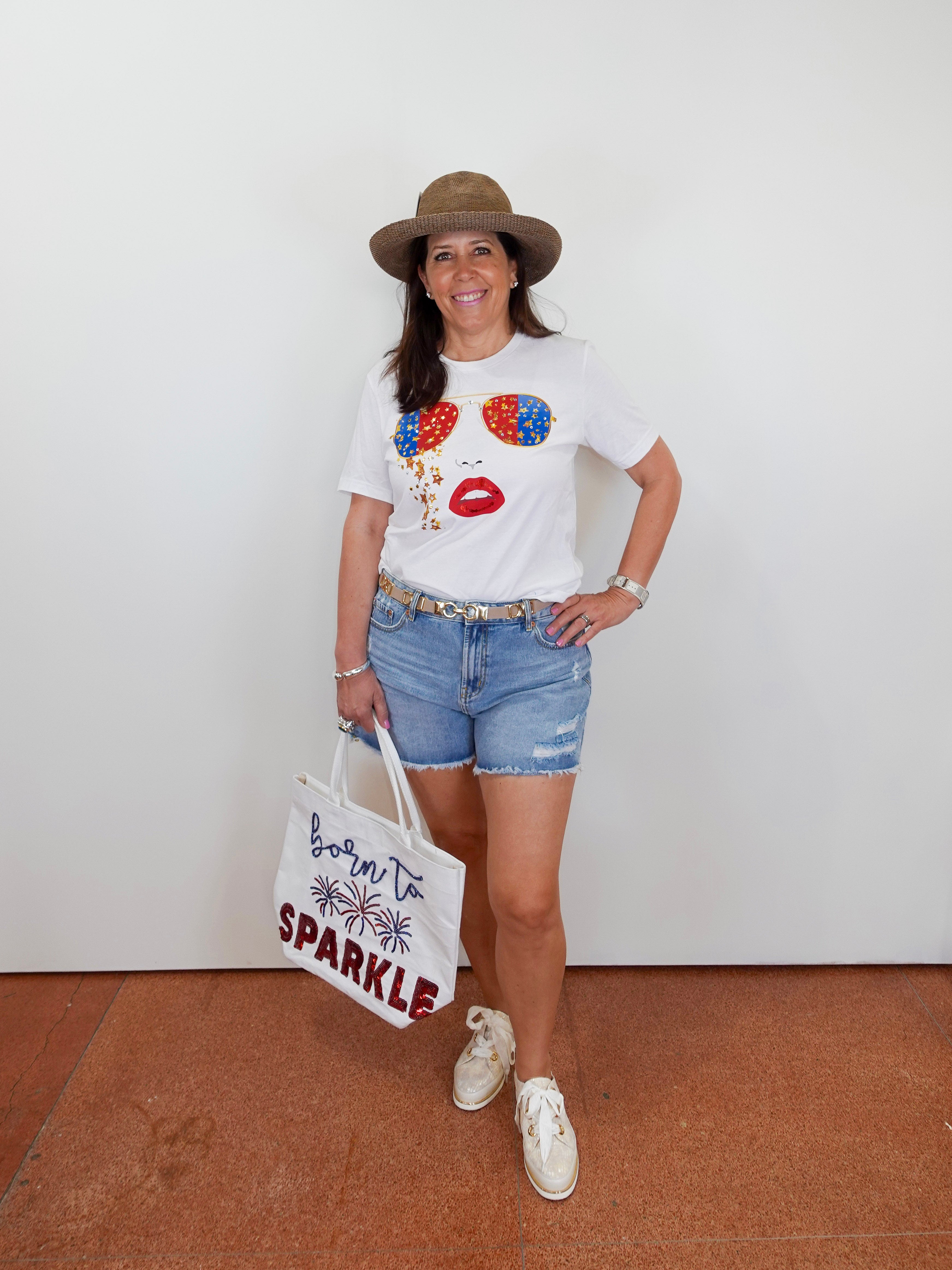 Woman wearing a white t-shirt with colorful design, denim shorts, and a hat, holding a tote bag.