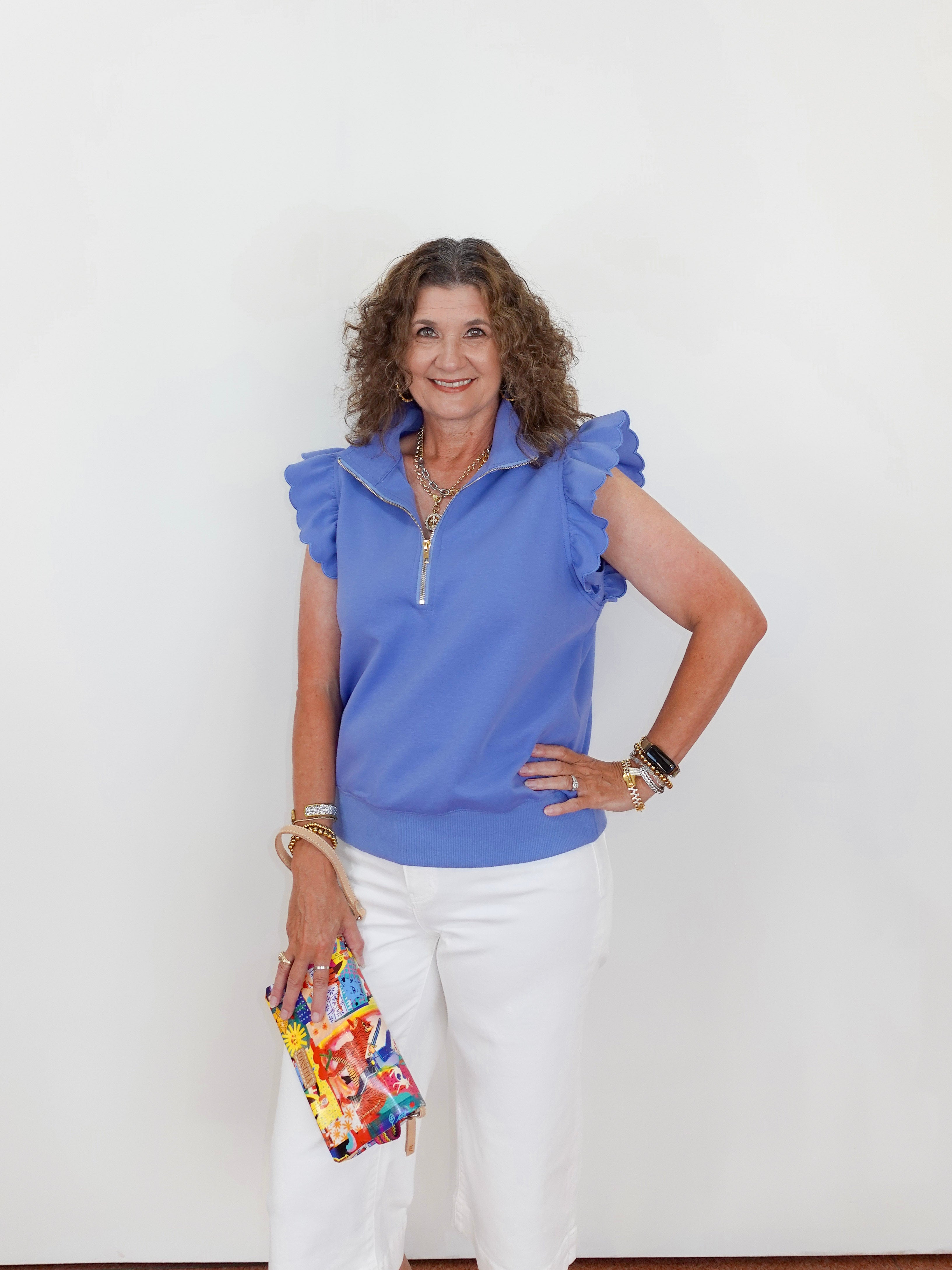 Woman wearing a blue top and white pants, holding a colorful clutch against a white background