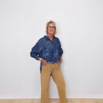 Person wearing a blue patterned shirt and beige pants against a white wall.