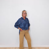 Person wearing a blue patterned shirt and beige pants against a white wall.