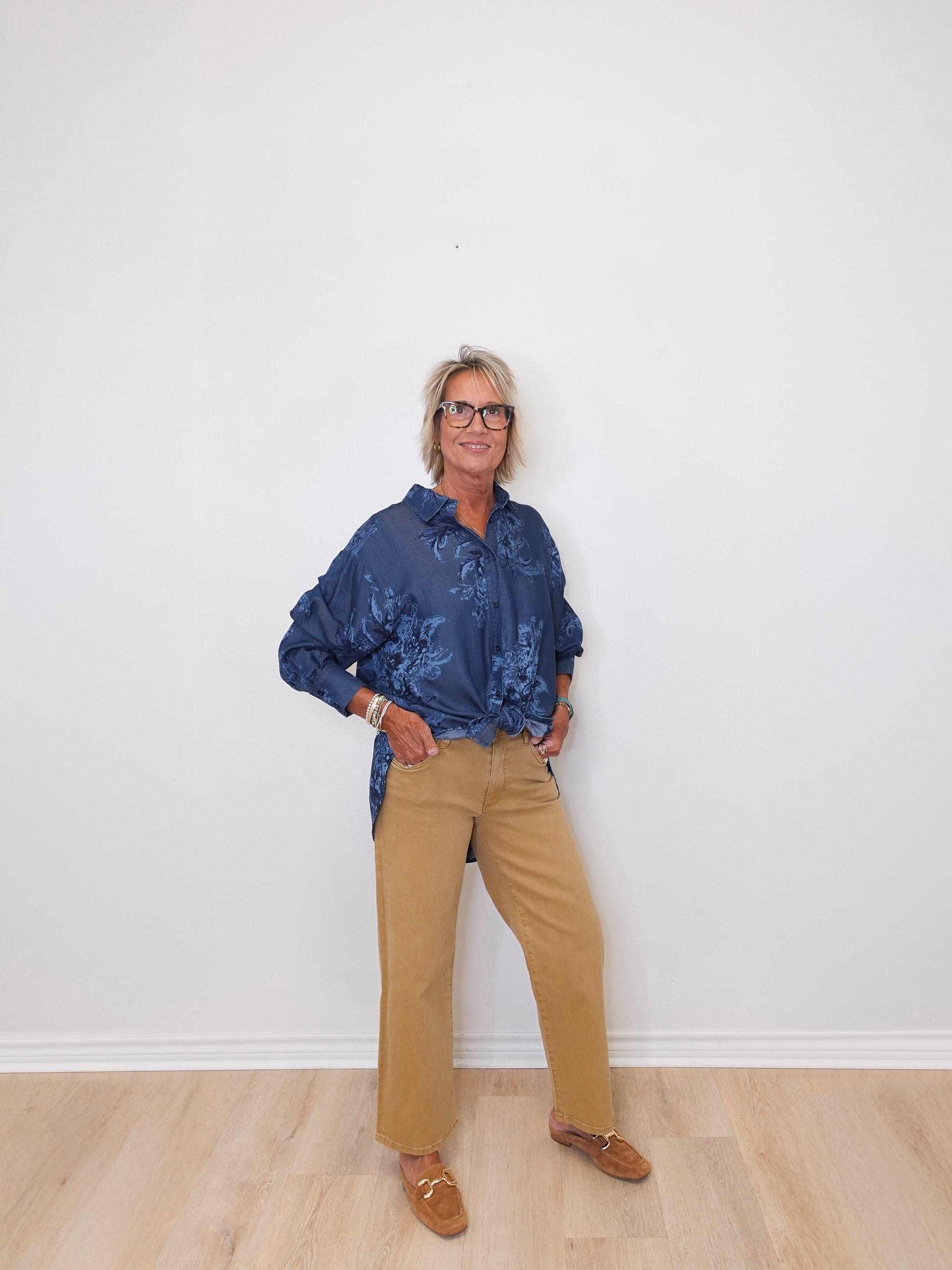 Person wearing a blue patterned shirt and beige pants against a white wall.