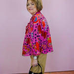 Woman wearing a colorful patterned jacket holding a black handbag with a white bow against a pink wall.