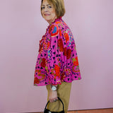 Woman wearing a colorful patterned jacket holding a black handbag with a white bow against a pink wall.