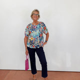 Woman wearing a colorful floral blouse and dark pants, holding a pink handbag against a white wall.