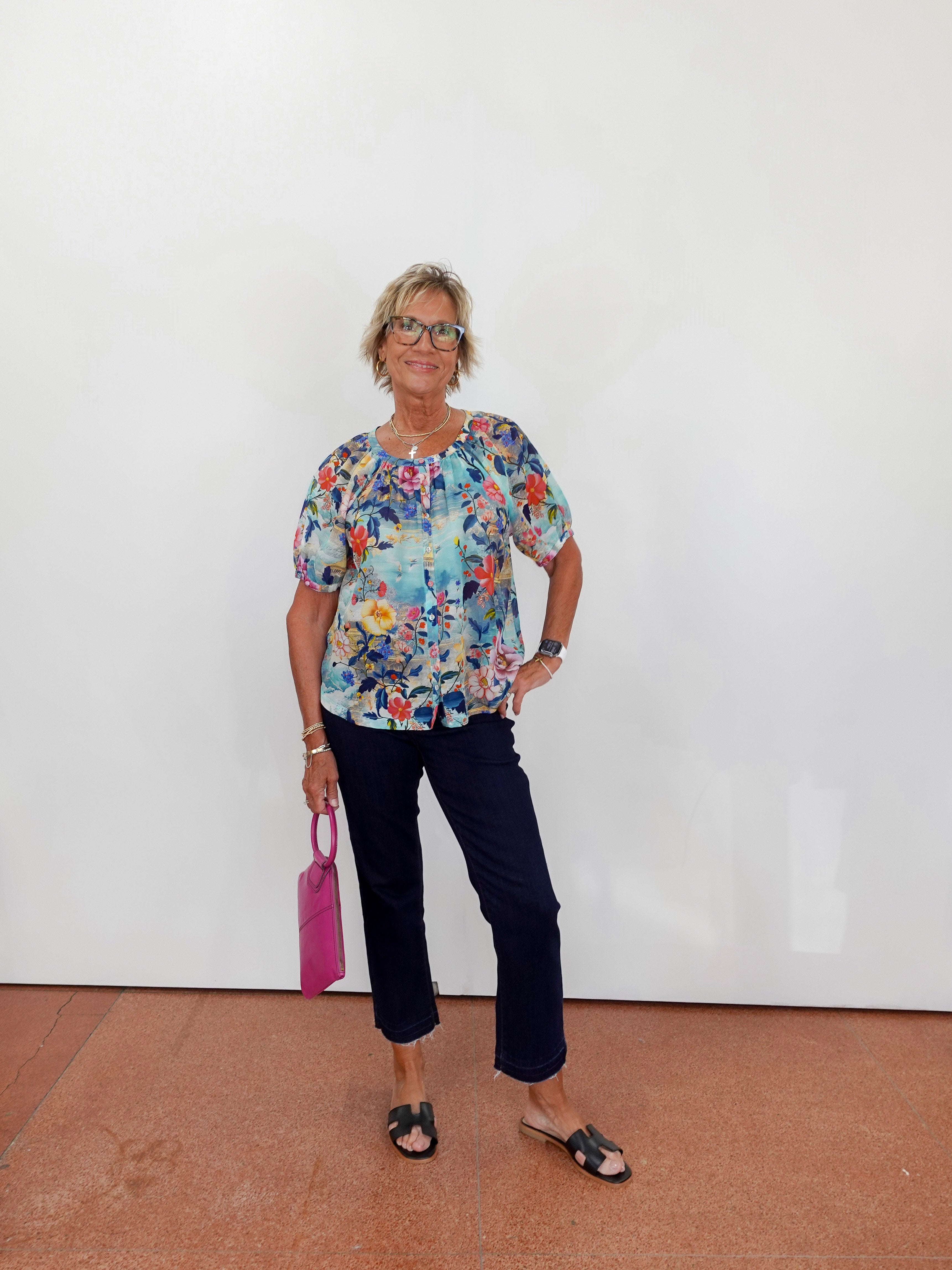 Woman wearing a colorful floral blouse and dark pants, holding a pink handbag against a white wall.