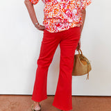 Person wearing a colorful floral blouse and red pants, holding a woven bag against a white wall.