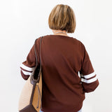 Person wearing a brown sweater with white stripes on sleeves, holding a beige handbag.