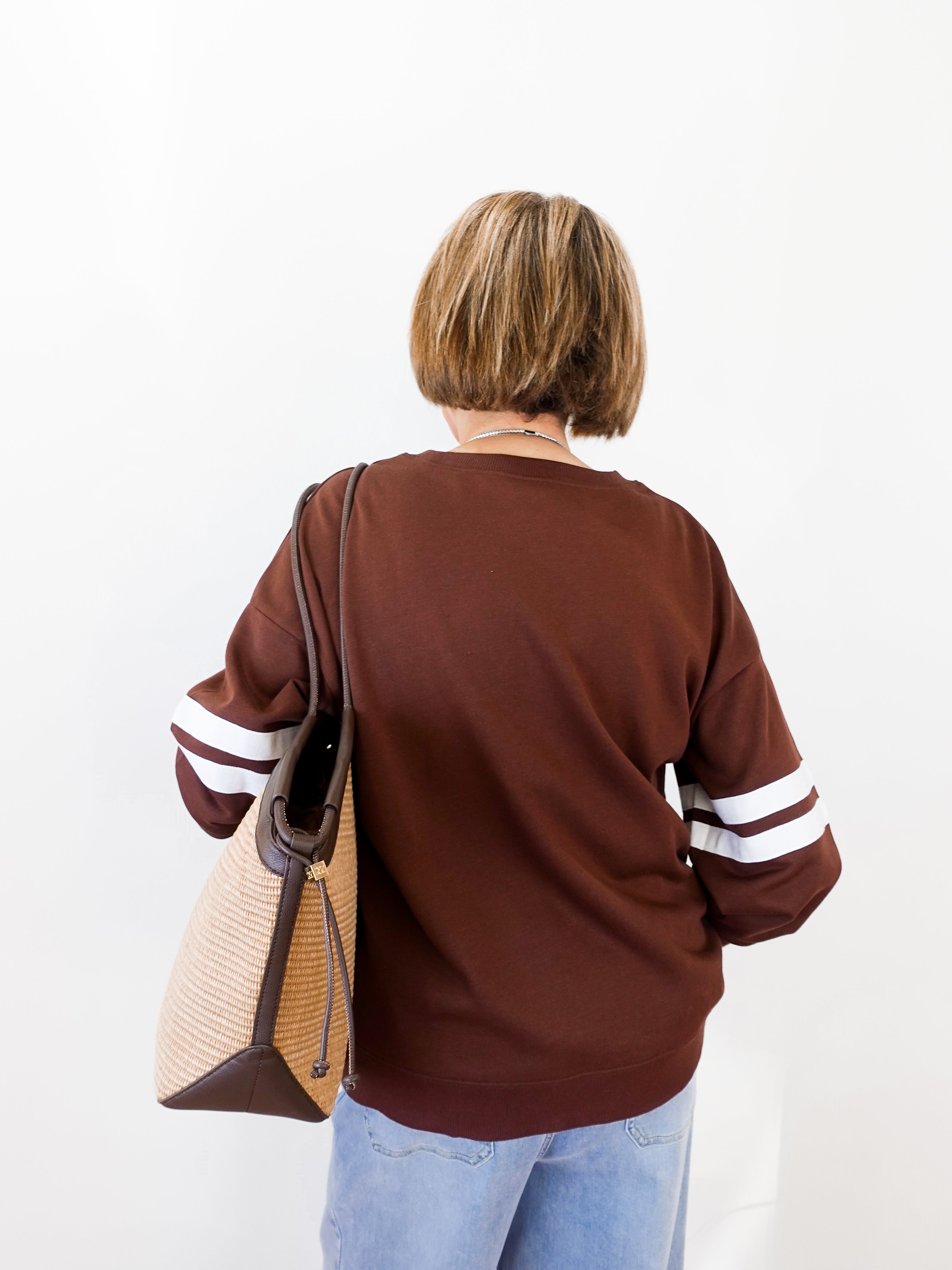 Person wearing a brown sweater with white stripes on sleeves, holding a beige handbag.