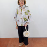 Woman wearing a floral blouse and black pants, holding a white handbag against a white wall.