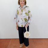 Woman wearing a floral blouse and black pants, holding a white handbag against a white wall.
