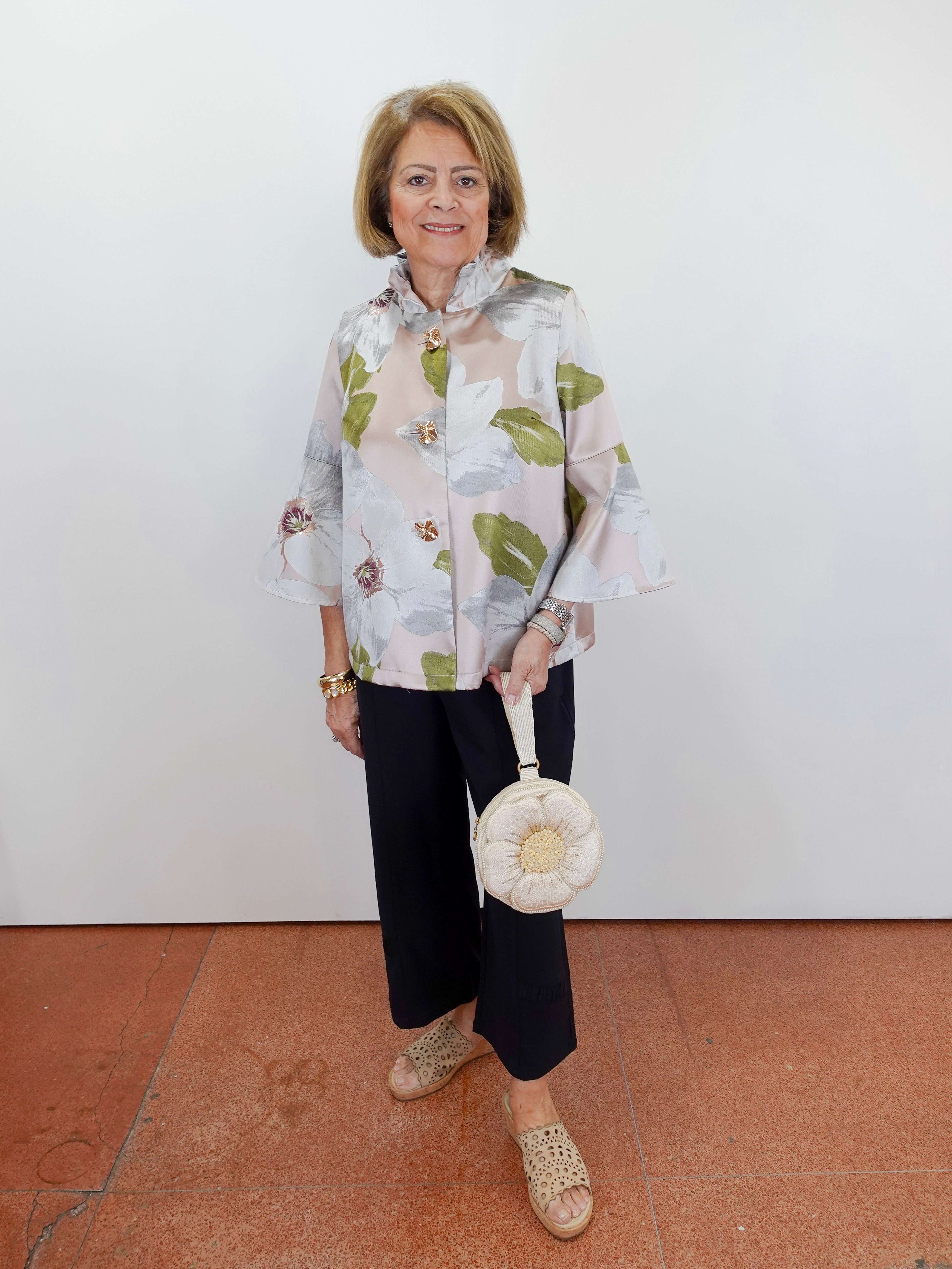 Woman wearing a floral blouse and black pants, holding a white handbag against a white wall.
