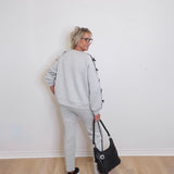 Person wearing a gray tracksuit holding a black handbag against a white wall.