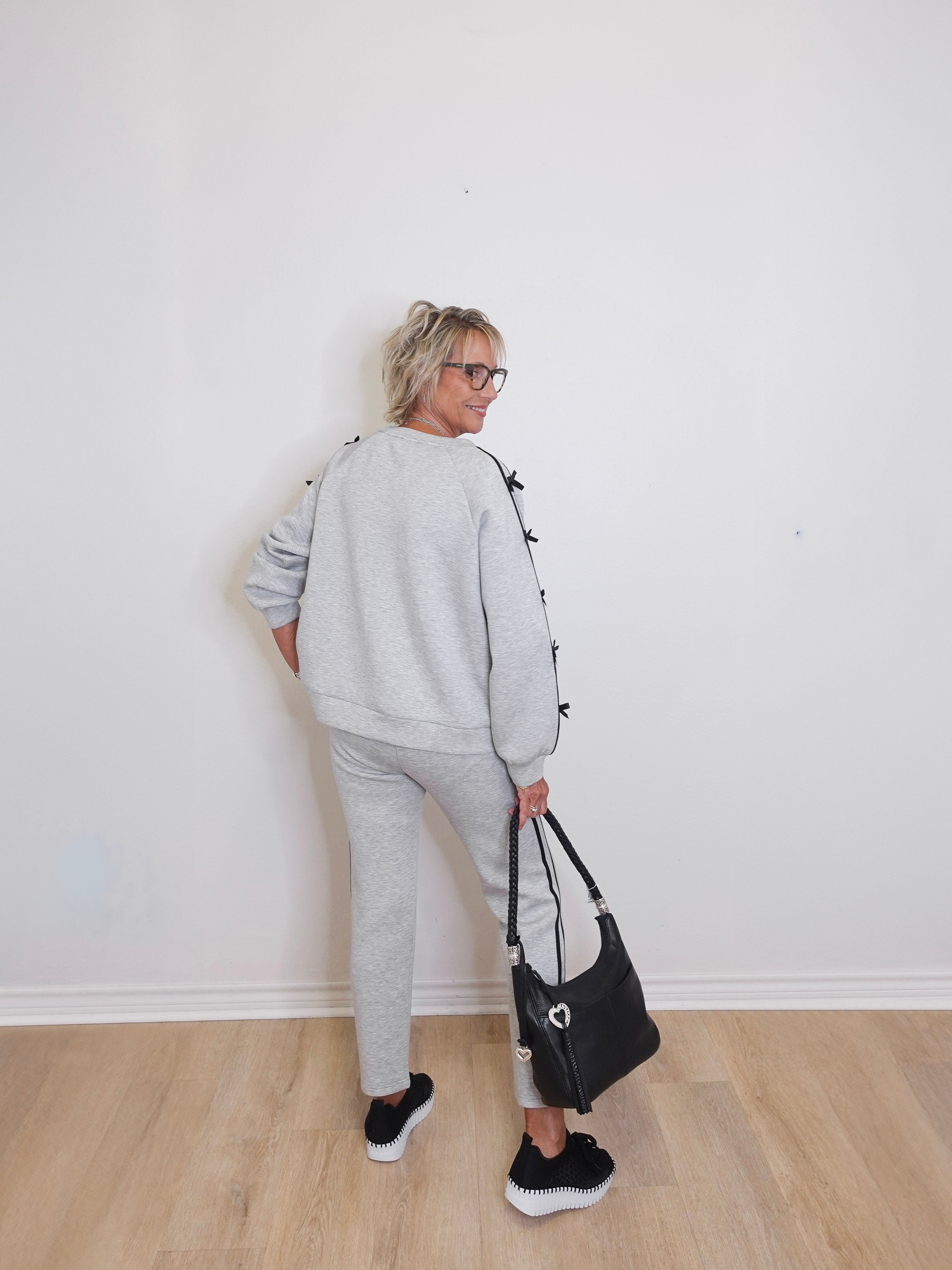 Person wearing a gray tracksuit holding a black handbag against a white wall.
