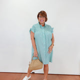 Woman wearing a light blue dress holding a beige handbag against a white wall.