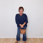 Woman wearing a navy blue top and jeans, holding a brown handbag against a white wall.