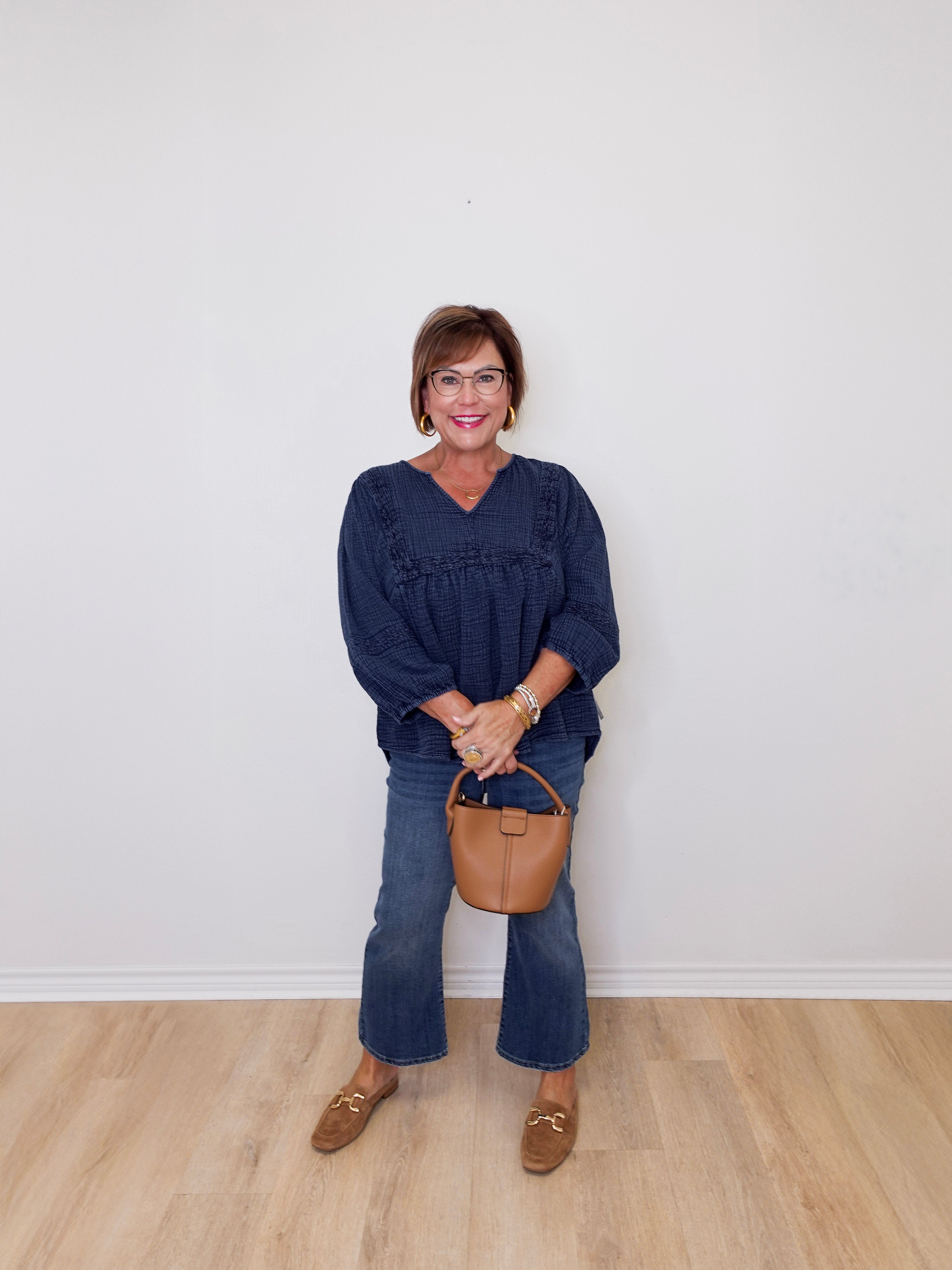 Woman wearing a navy blue top and jeans, holding a brown handbag against a white wall.