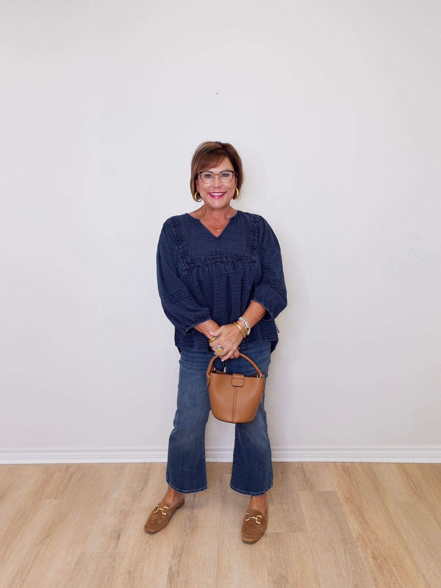Woman wearing a navy blue top and jeans, holding a brown handbag against a white wall.