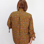A woman viewed from behind wearing a brown long-sleeve button-up shirt with a colorful geometric pattern, paired with blue jeans and holding a small red handbag against a plain white background.