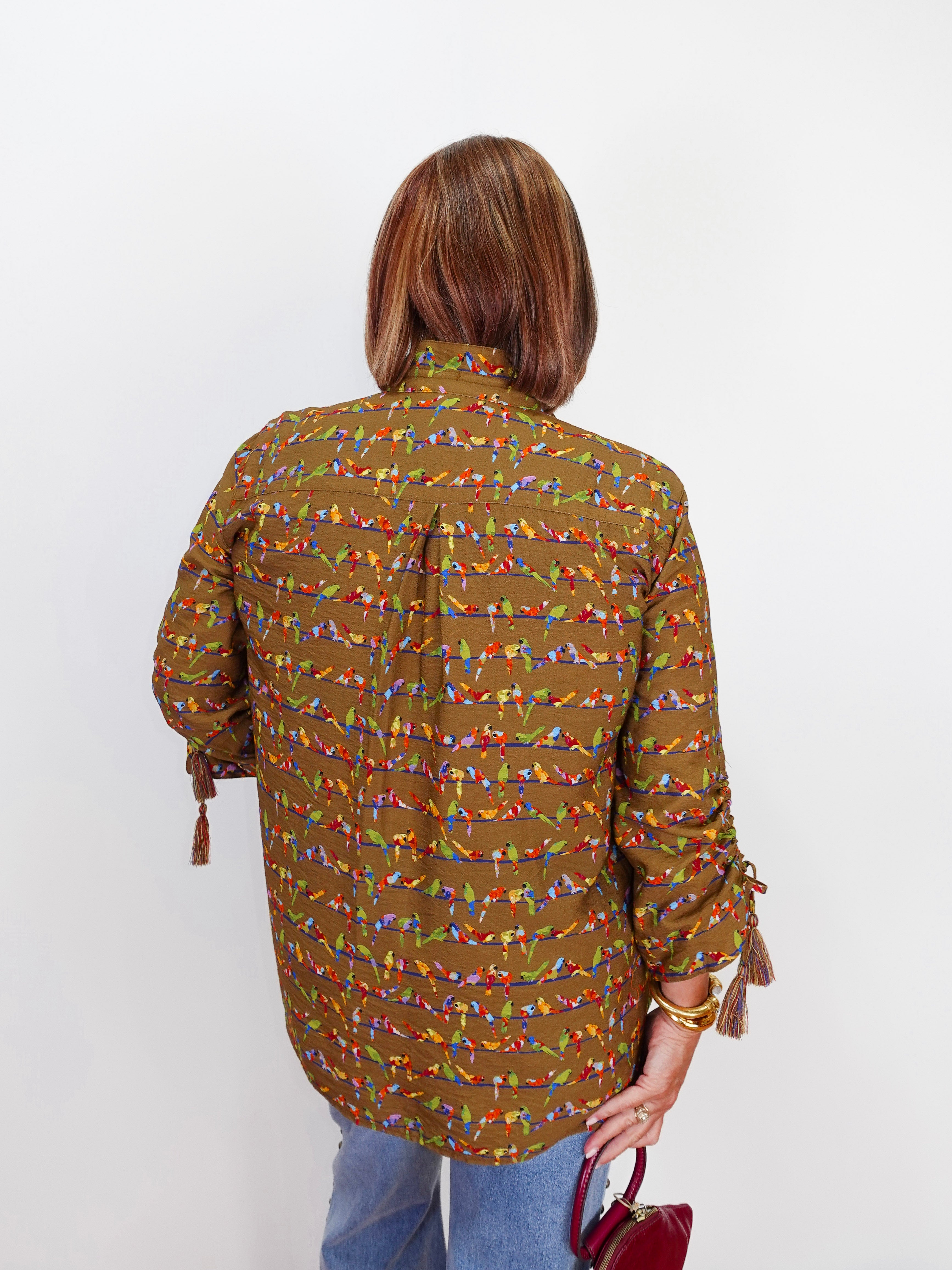 A woman viewed from behind wearing a brown long-sleeve button-up shirt with a colorful geometric pattern, paired with blue jeans and holding a small red handbag against a plain white background.