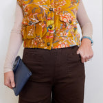 Person wearing a patterned mustard yellow vest over a beige long-sleeve shirt, holding a blue clutch.