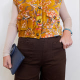 Person wearing a patterned mustard yellow vest over a beige long-sleeve shirt, holding a blue clutch.