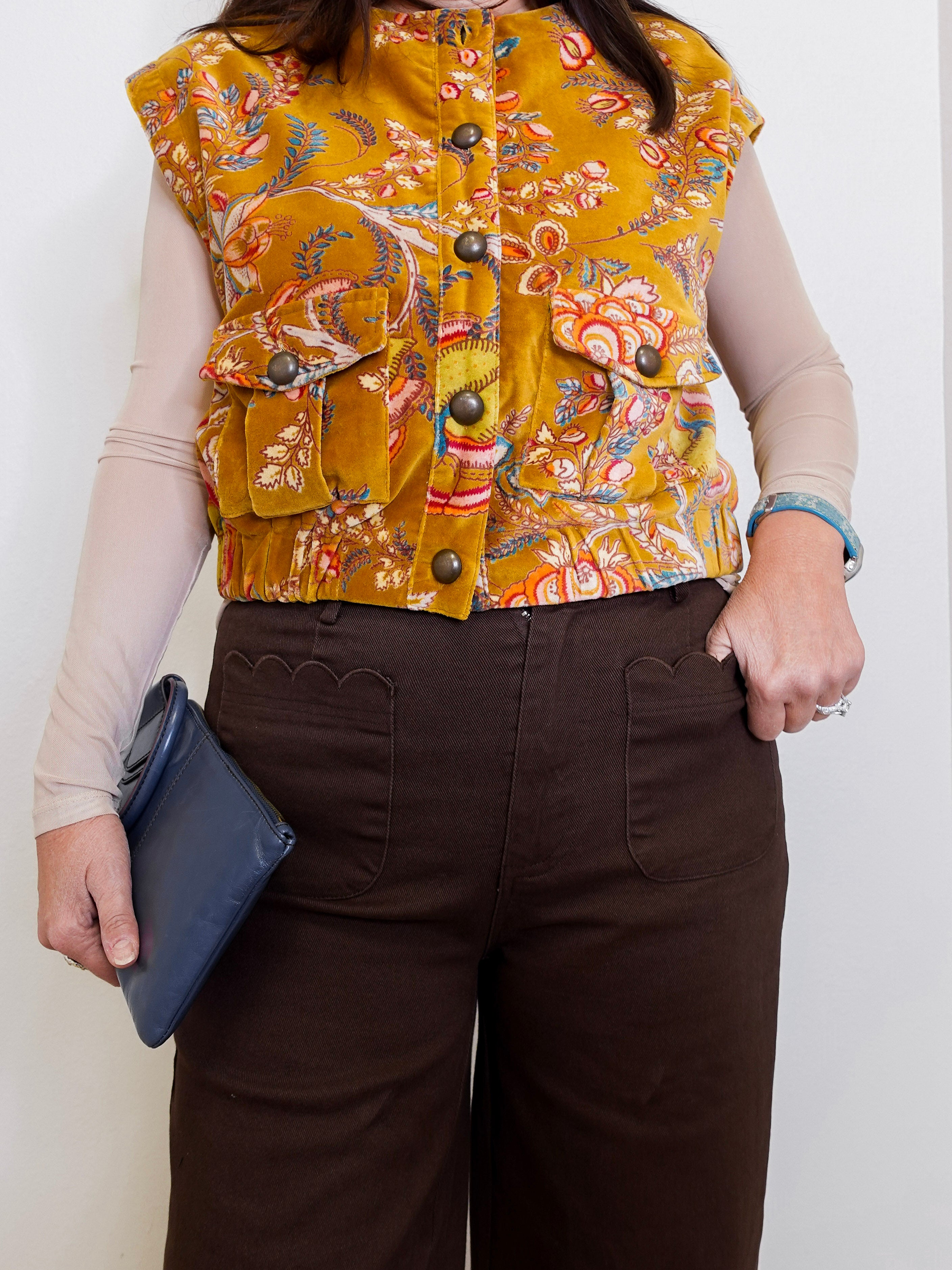 Person wearing a patterned mustard yellow vest over a beige long-sleeve shirt, holding a blue clutch.
