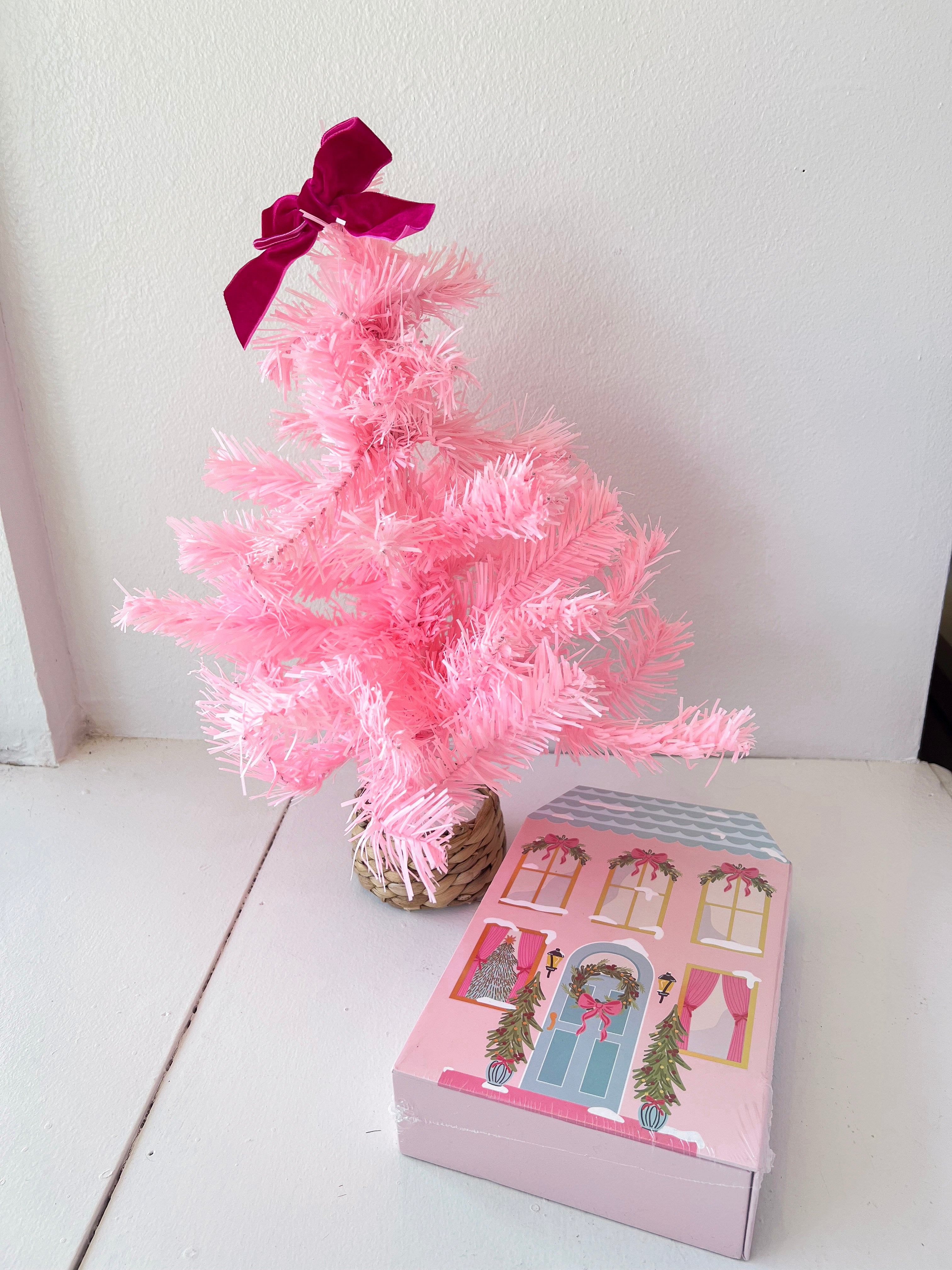 Small pink artificial Christmas tree with a purple bow next to a decorative box on a white surface.