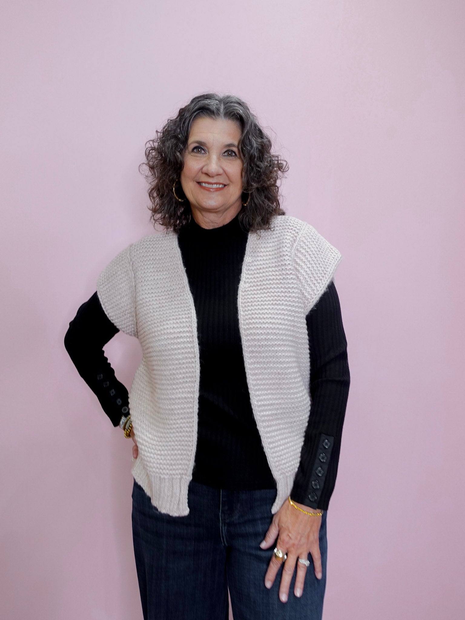 Woman wearing a white knit vest over a black long-sleeve shirt on a pink background
