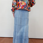 Person wearing a colorful floral jacket and blue tiered skirt on a plain background