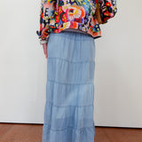 Person wearing a colorful floral jacket and blue tiered skirt on a plain background