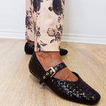 Black woven flats with a gold buckle worn with floral pants on a wooden floor.