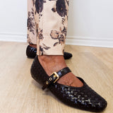 Black woven flats with a gold buckle worn with floral pants on a wooden floor.