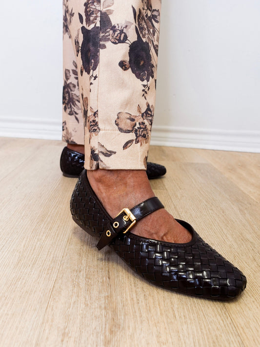 Black woven flats with a gold buckle worn with floral pants on a wooden floor.