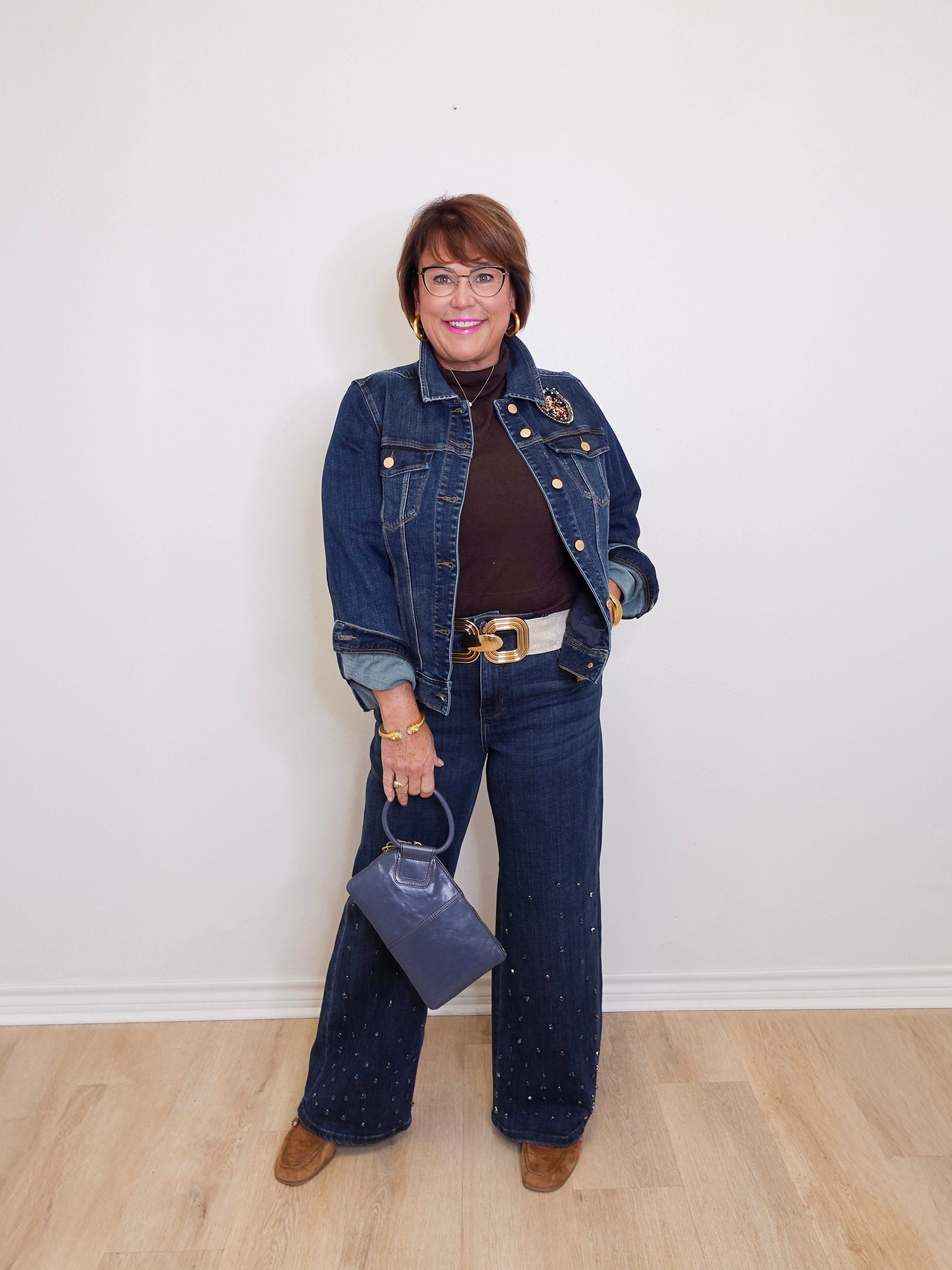 Person wearing a denim jacket and jeans holding a handbag against a white wall.