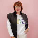 Woman wearing a white sweatshirt with colorful letters, black vest, and white pants against a pink background