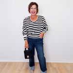 Woman wearing a black and white striped sweater and blue jeans, standing against a white wall.