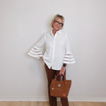 Woman wearing a white blouse with bell sleeves, brown pants, and holding a brown handbag against a plain background.