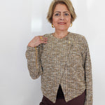 An older woman with short blonde hair models a textured tweed jacket with gold buttons and a gold zipper detail on the sleeve, paired with dark brown trousers, against a plain white background.