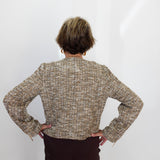 A person viewed from behind wearing a textured tweed jacket in beige, brown, and gray tones, paired with dark brown trousers, standing against a plain white wall.