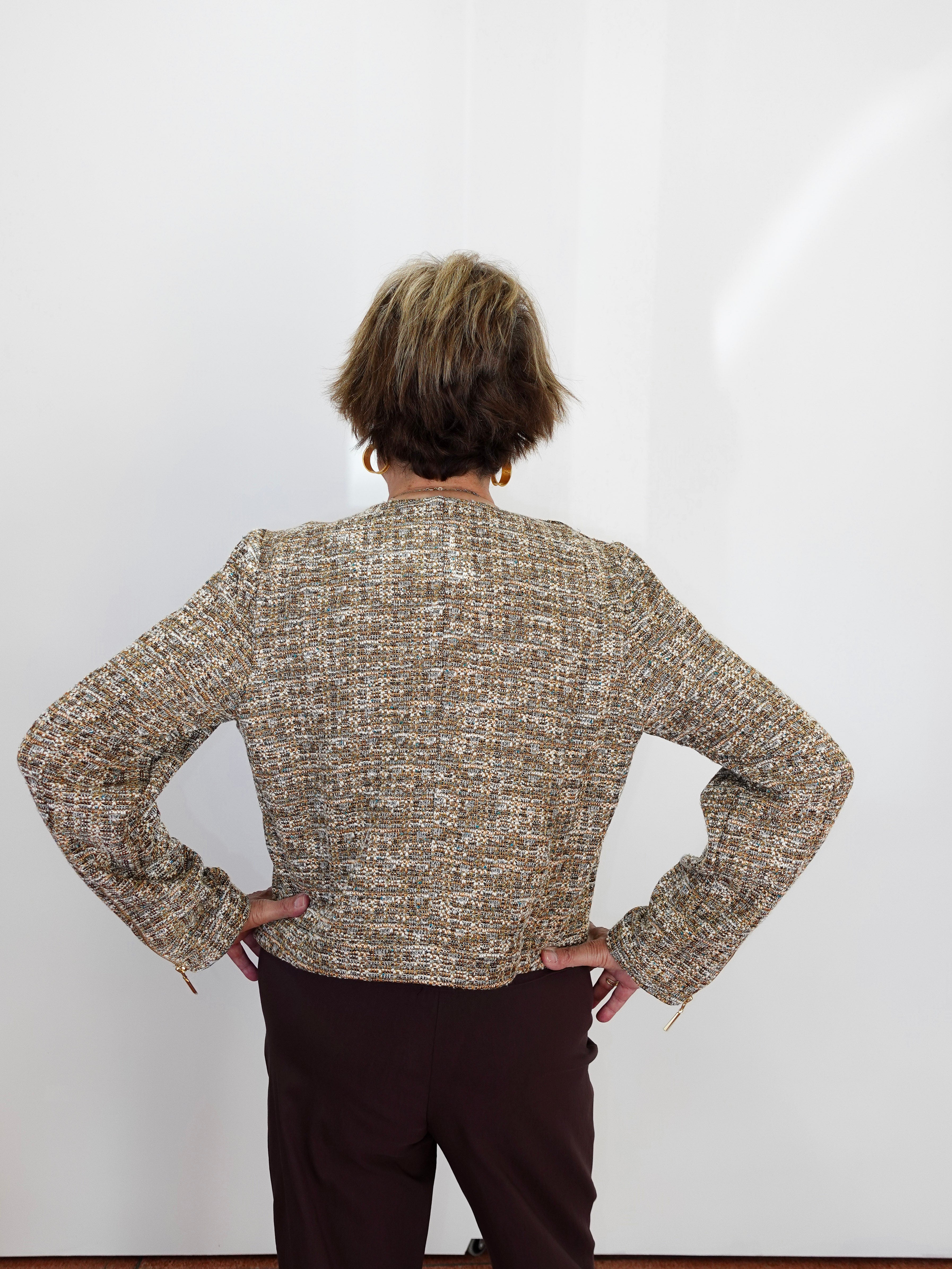 A person viewed from behind wearing a textured tweed jacket in beige, brown, and gray tones, paired with dark brown trousers, standing against a plain white wall.