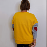 Person wearing a yellow sweater with gray sleeves and red elbow patches, standing against a white wall.