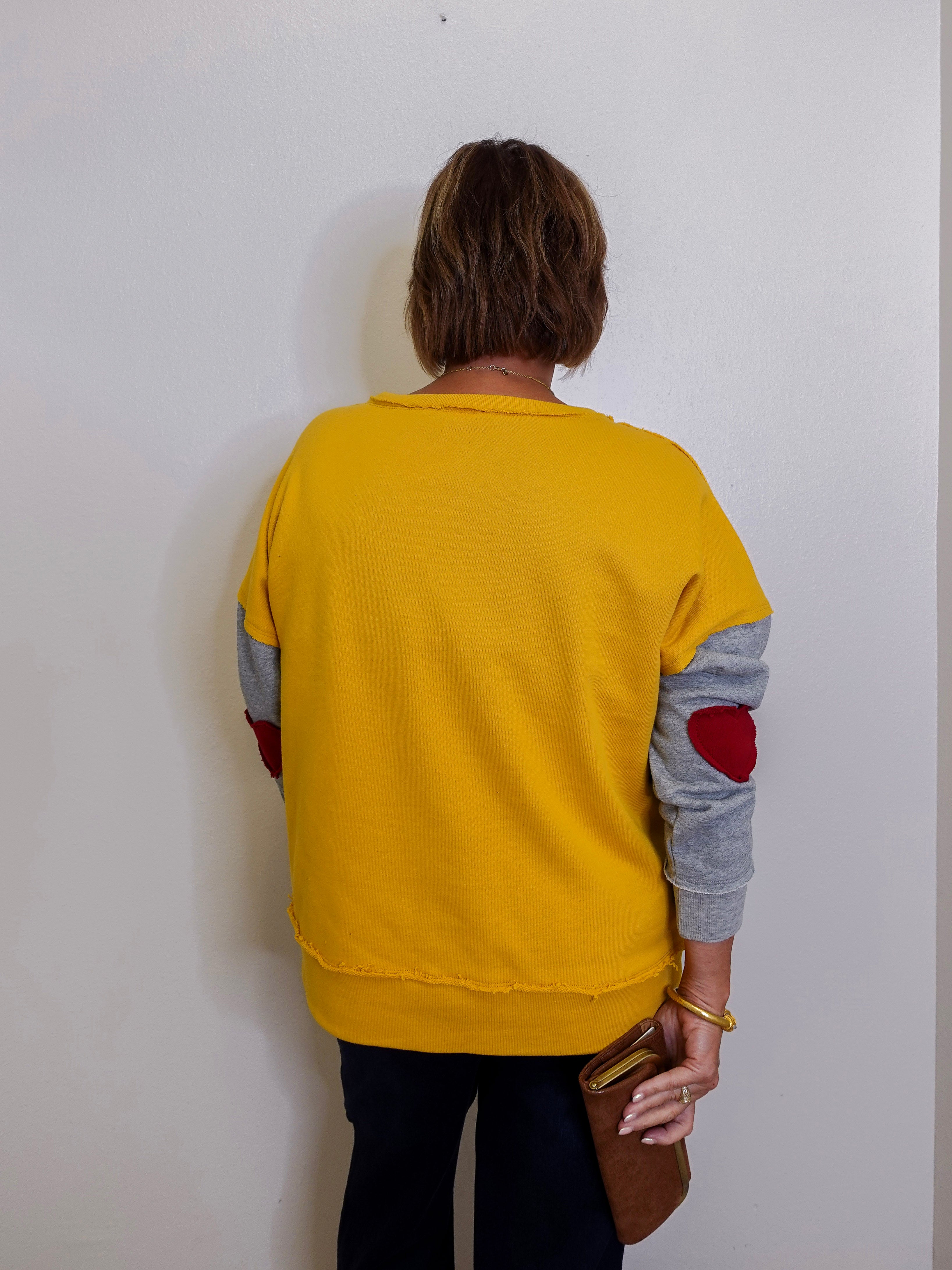 Person wearing a yellow sweater with gray sleeves and red elbow patches, standing against a white wall.