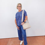 Woman wearing a blue patterned top and jeans, holding a beige handbag against a white wall.