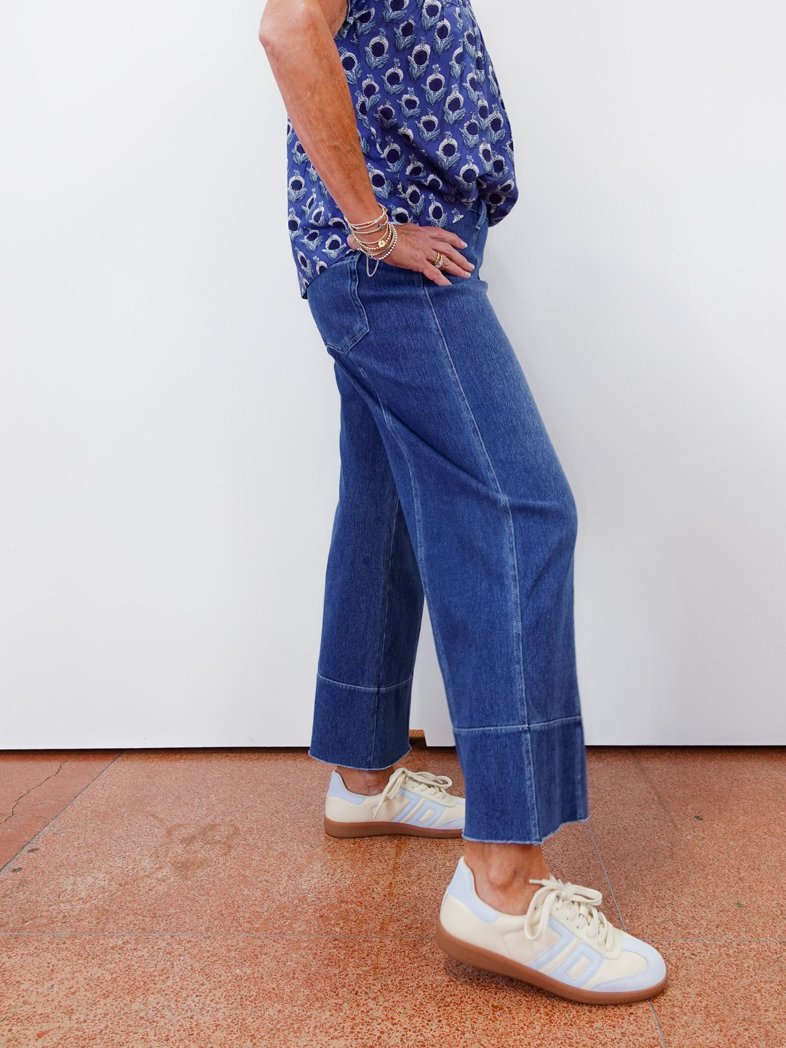 Person wearing a blue patterned top and blue jeans with white sneakers on a plain background