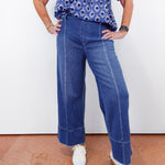 Person wearing a blue patterned shirt and blue jeans against a white wall.
