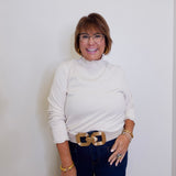 Woman wearing a white turtleneck sweater and blue jeans with a gold belt against a white background