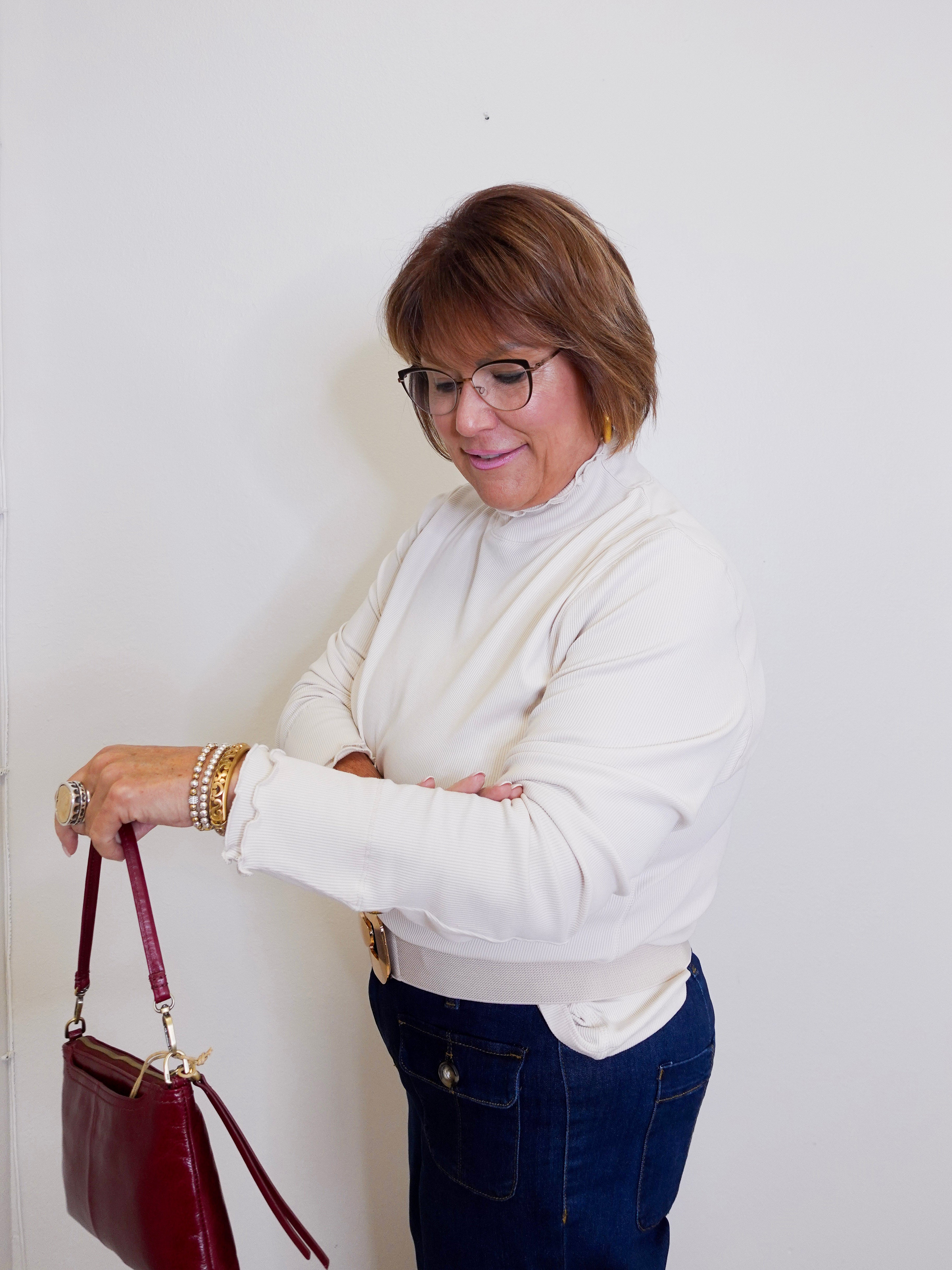 Woman in a white sweater and blue jeans holding a red handbag against a white wall.