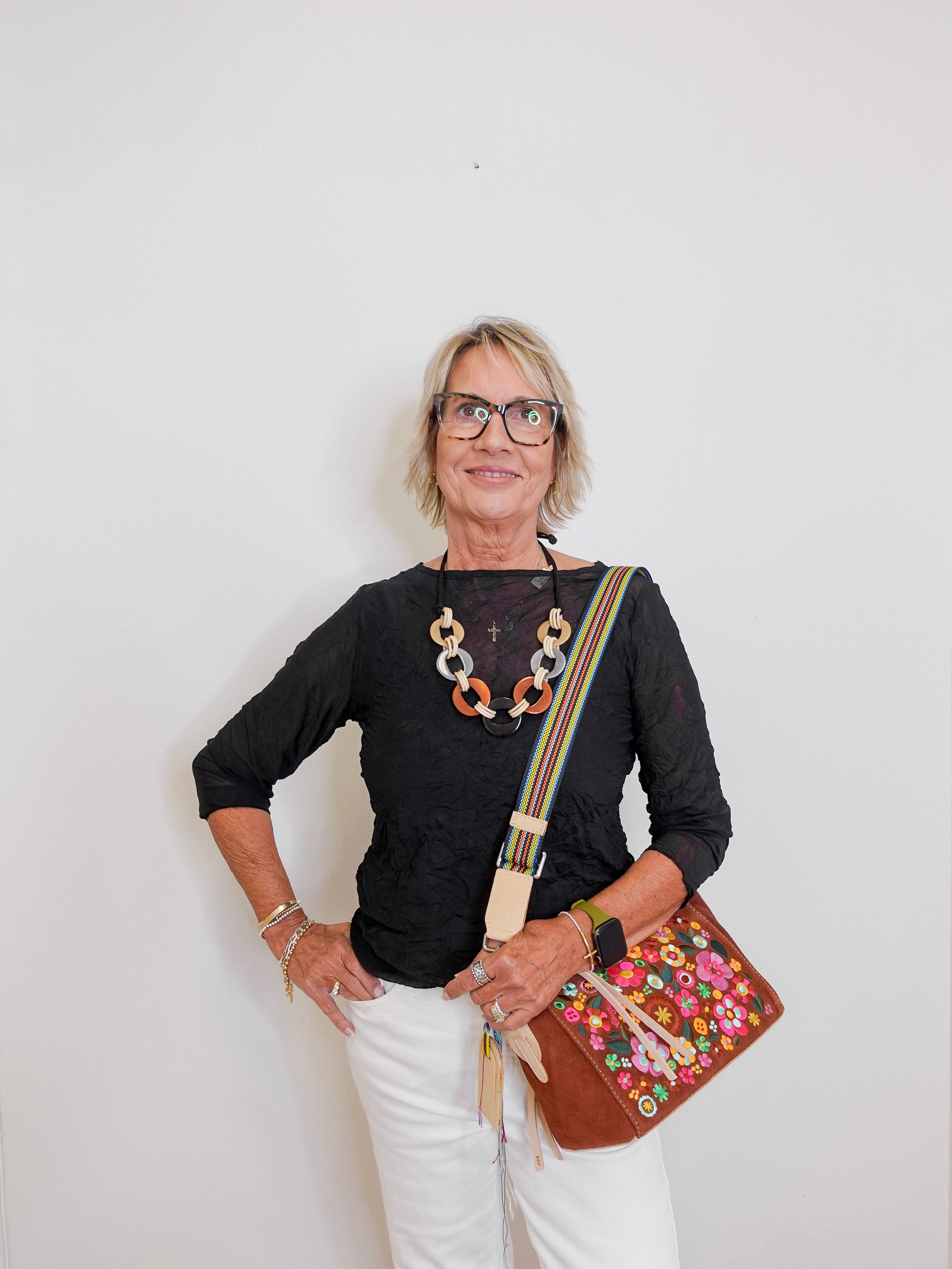 Woman wearing a black top, white pants, and carrying a colorful handbag against a plain background