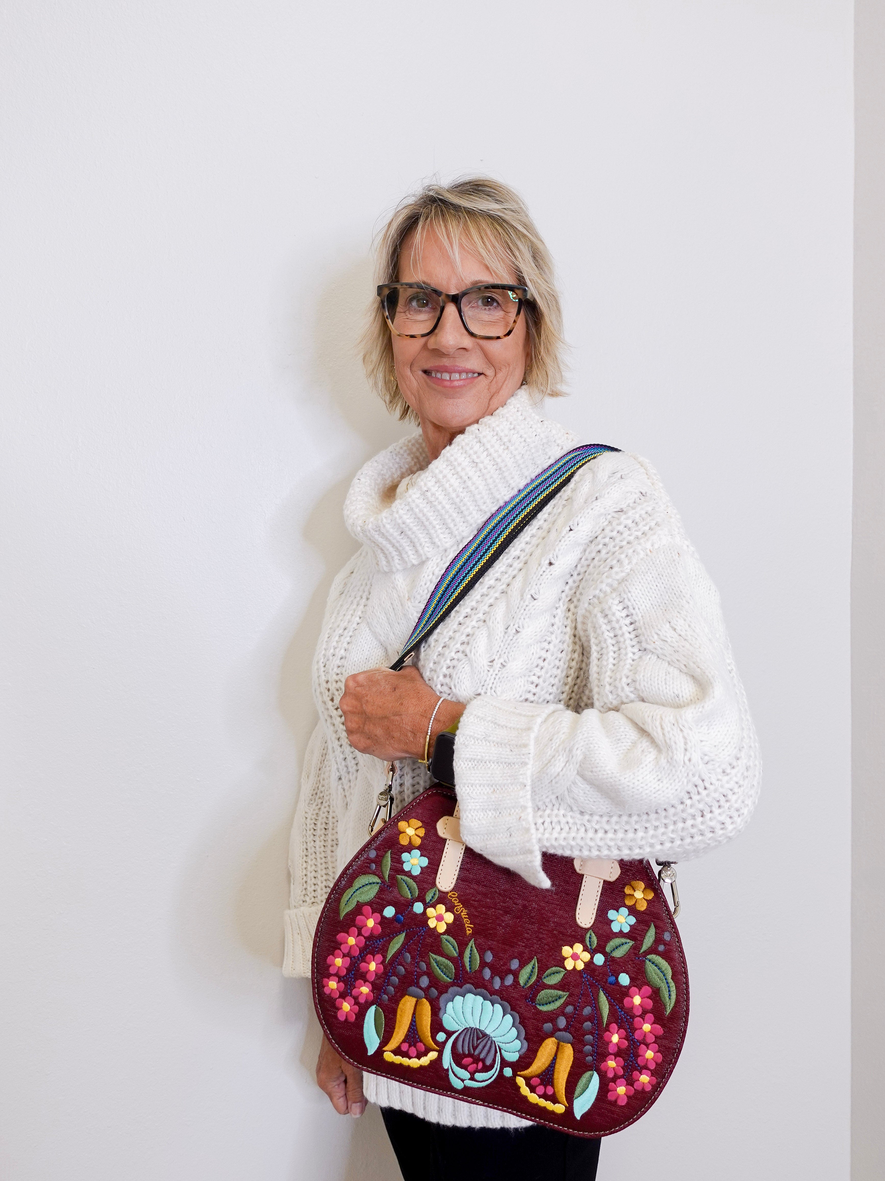 Woman wearing a white knitted sweater holding a colorful embroidered bag against a plain background